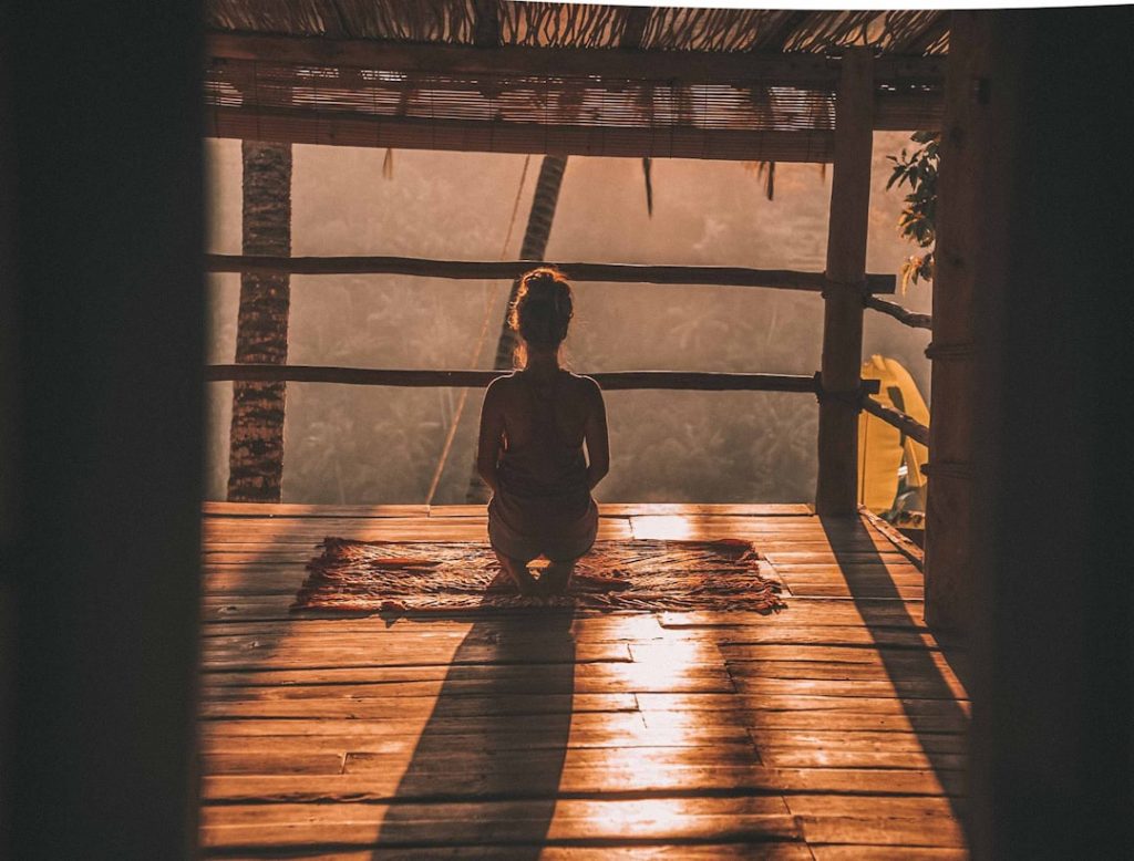 A woman sitting in meditation on a wooden floor, looking out over trees in soft morning light.