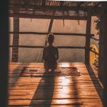 A woman sitting in meditation on a wooden floor, looking out over trees in soft morning light.