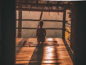 A woman sitting in meditation on a wooden floor, looking out over trees in soft morning light.