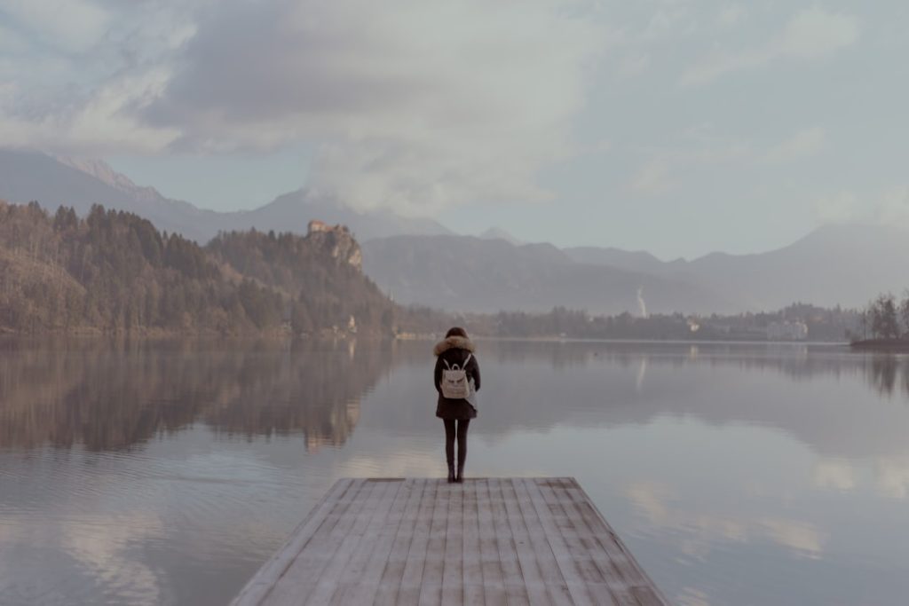 A lone figure standing beside calm water in a quiet landscape.
