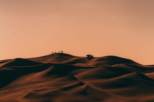 Silhouettes of travellers moving across desert sand at sunset.